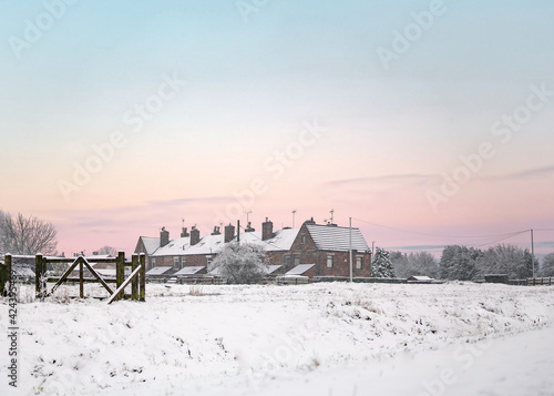 Beautiful winter sunset over row of old houses in countryside with deep snow and glowing sky. Snowy scene as sunrise at dawn pink orange skies. Gate and wooden fence in foreground.