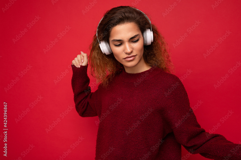 Fototapeta premium Photo of beautiful happy smiling young brunette curly woman wearing dark red sweater isolated over red background wall wearing white bluetooth headphones listening to music and having fun looking to
