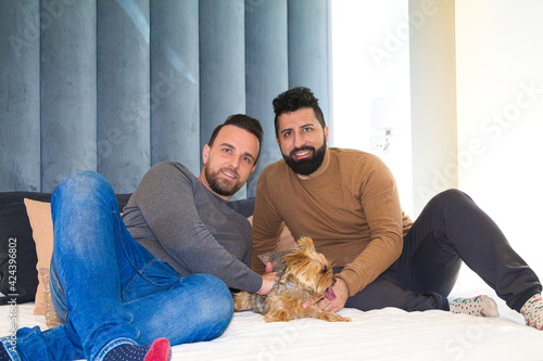 gay couple sitting on the bed playing with their pet. They are looking at camera happy.