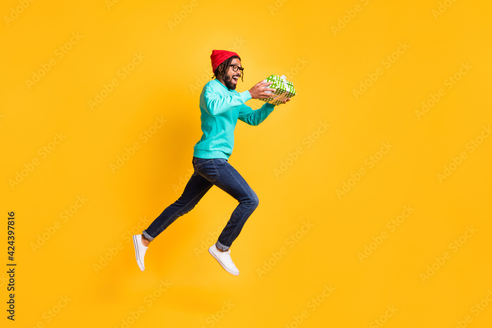 Full size profile photo of brunette optimistic guy jump run take present wear cap spectacles pullover jeans isolated on yellow background