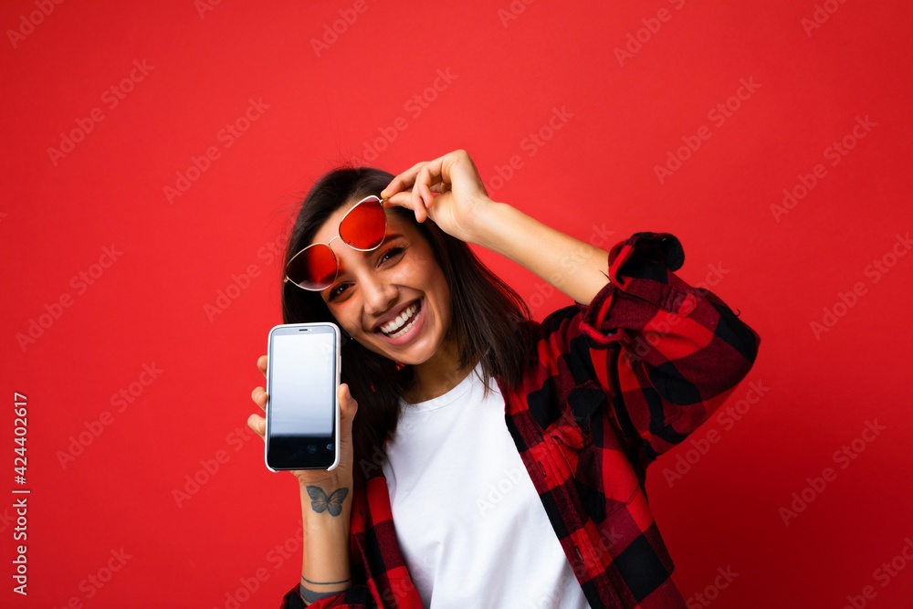 Photo of beautiful smiling happy young woman good looking wearing casual stylish red shirt white t-shirt and red sunglasses standing isolated on red background with copy space holding smartphone Photo of beautiful smiling happy young woman good looking wearing casual stylish red shirt white t-shirt and red sunglasses standing isolated on red background with copy space holding smartphone