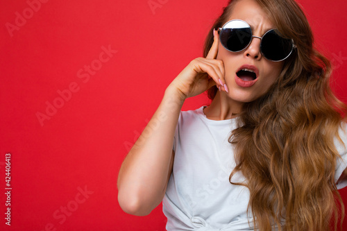 Closeup shot of beautiful surprised young dark blonde curly woman isolated over red background wall wearing casual white t-shirt and stylish sunglasses looking at camera with open mouth