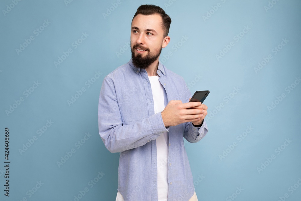 Photo shot of handsome positive good looking young man wearing casual stylish outfit poising isolated on background with empty space holding in hand and using mobile phone messaging sms looking to the Photo shot of handsome positive good looking young man wearing casual stylish outfit poising isolated on background with empty space holding in hand and using mobile phone messaging sms looking to the