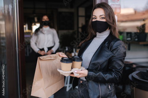 Attractive female customer in face mask standing outdoors by cafe and take away food and coffee. Background of cashier in mask waiting for next customer. New norm during quarantine. Covid19
