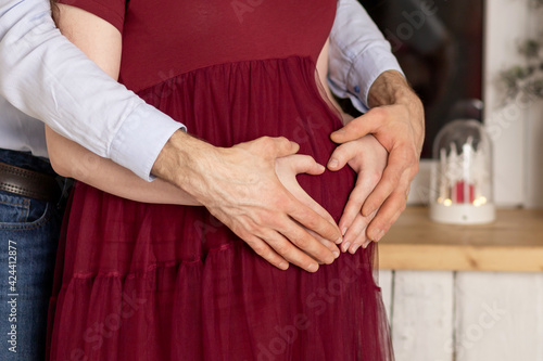 Couple holding hands in the shape of heart on pregnant belly. Young parents expecting baby, child, kid. Happy family. Pregnancy mom. Closeup of man hugging his pregnant wife for big tummy