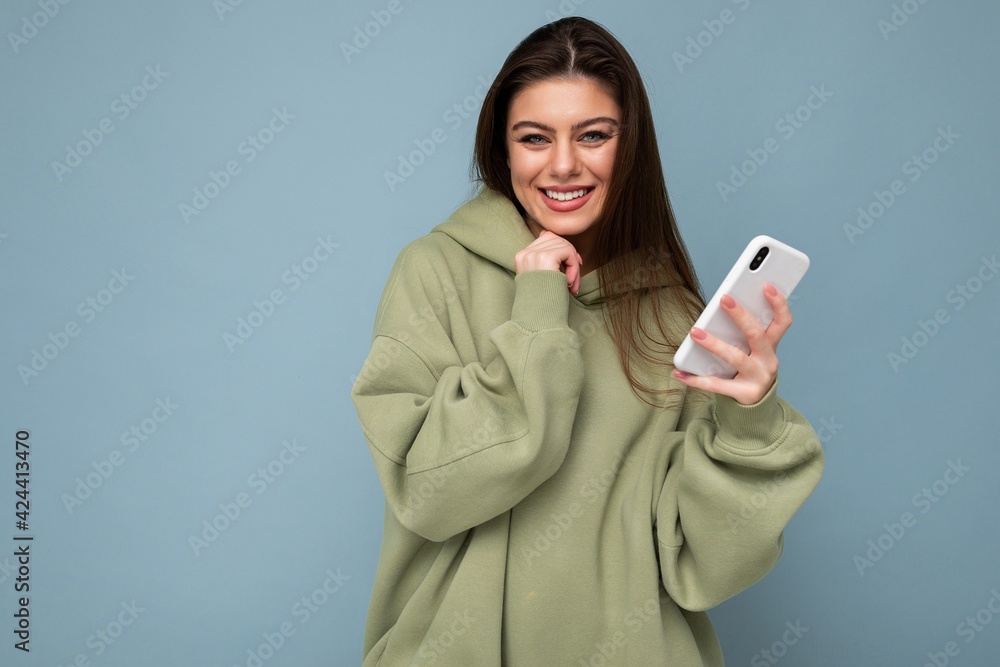 Photo of smiling sexy Beautiful young brunette woman wearing stylish green hoodie using mobile phone writing message isolated on background looking at camera