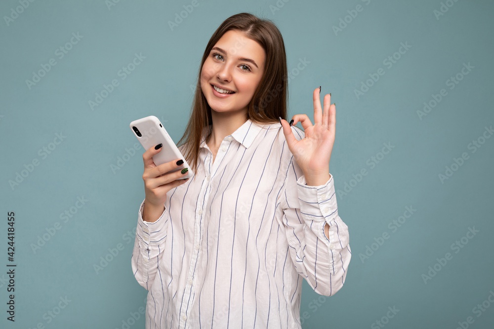 Photo of smiling beautiful young blonde woman wearing casual white shirt isolated over blue background wall holding smartphone and texting message looking at camera and showing OK gesture Photo of smiling beautiful young blonde woman wearing casual white shirt isolated over blue background wall holding smartphone and texting message looking at camera and showing OK gesture