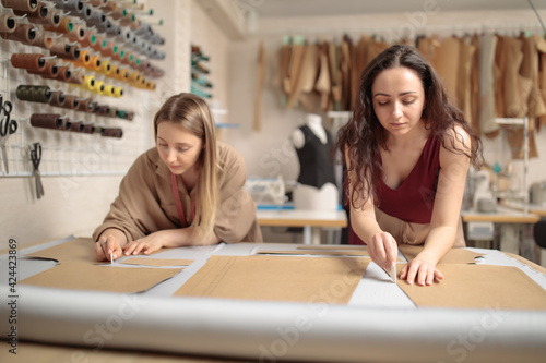 portrait of two young beautiful women tailors or fashion designers working with clothing patterns on workshop studio while sewing classical pants in atelier