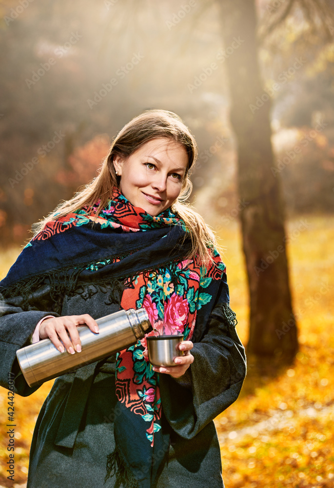 Attractive smiling woman with thermos of tea in autumn park on sunny day