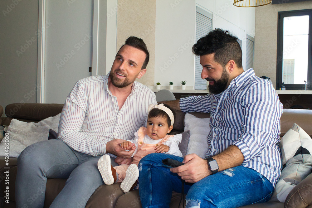 gay couple and their daughter sitting on the sofa watching television. The girl watches the cartoons attentively. gay couple and their daughter sitting on the sofa watching television. The girl watches the cartoons attentively.