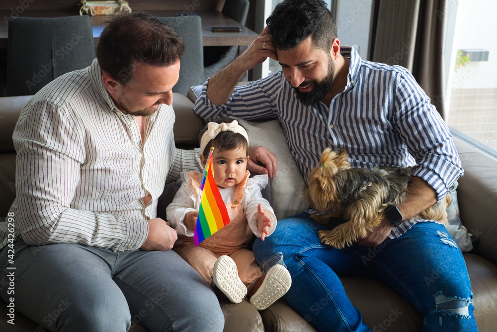 gay couple with their young daughter and dog sitting on the sofa watching television. The little girl watches the cartoons attentively and holds the gay pride flag in her hand.