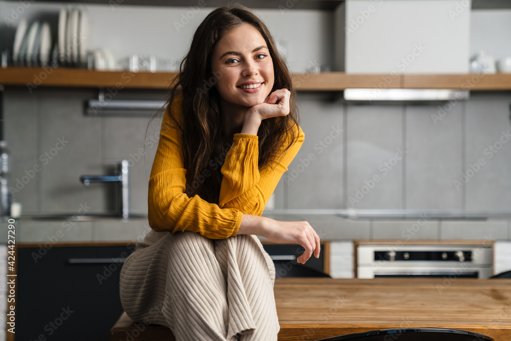 Young brunette woman smiling at camera while sitting on table in kitchen Young brunette woman smiling at camera while sitting on table in kitchen
