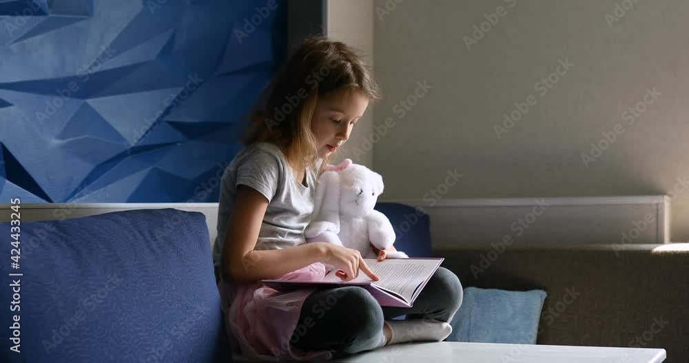 Cute little girl eading a book with stuffed teddy bunny toy and smiling sitting on the sofa in the living room. Stay at home during coronavirus covid-19 pandemic Cute little girl eading a book with stuffed teddy bunny toy and smiling sitting on the sofa in the living room. Stay at home during coronavirus covid-19 pandemic