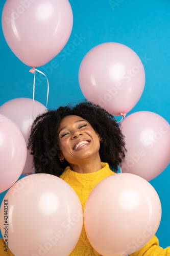 Happy pleased African woman with curly hair holding a lot of pink balloons, enjoys cool party, wears yellow sweater, celebrates birthday, looking up standing over studio blue background. Festive event