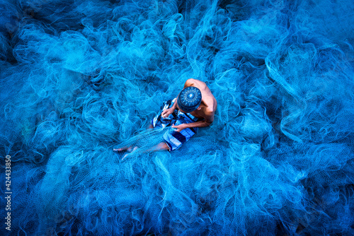 Old fisherman hands sewing blue fishing nets sitting on the ground and surrounded big net photo