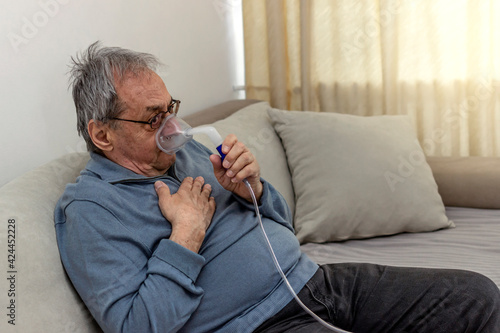 Grey haired senior man doing inhalation through oxygen mask at home. Inhalation treatment on senior man by the mask of inhaler during coronavirus quarantine. Senior male receiving oxygen therapy.