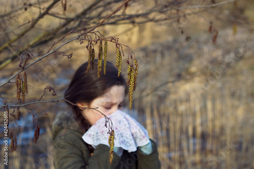 Unfocused caucasian girl with dark hairs is keeping a handkerchief near her nose because of seasonal allergy under the blossoming twigs. Alder tree pollen. Spring trees blooming. 