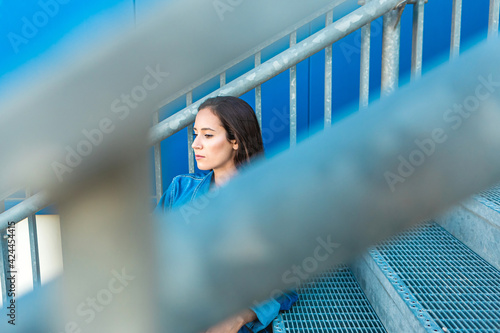 Lifestyle portrait of a young and beautiful woman sitting in a metallic stairs in the city in blue background