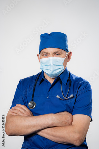 Portrait of happy senior doctor with folded arms and mask on face isolated on white background. Confident male doctor in a blue labcoat and stethoscope looking at camera. Portrait of handsome mature