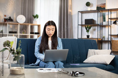 Young asian girl student sitting on couch at living room and working on wireless laptop. Attractive female student using portable computer for work and study remotely at home.