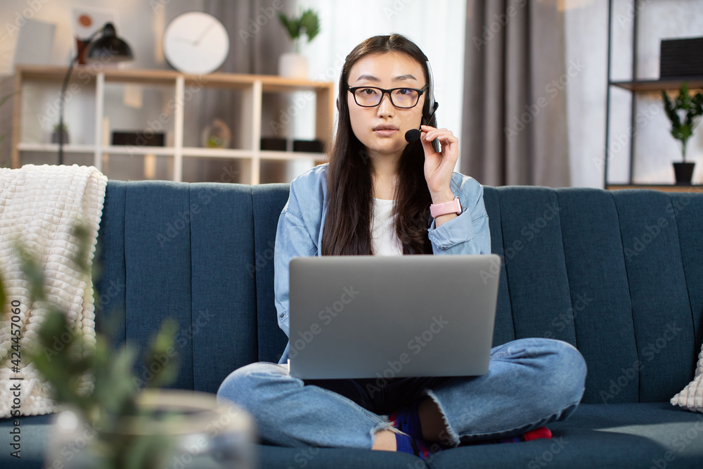 Serious asian woman using headset and laptop for video conference on laptop. Young female in eyeglasses sitting on couch and talking throught video call. Serious asian woman using headset and laptop for video conference on laptop. Young female in eyeglasses sitting on couch and talking throught video call.