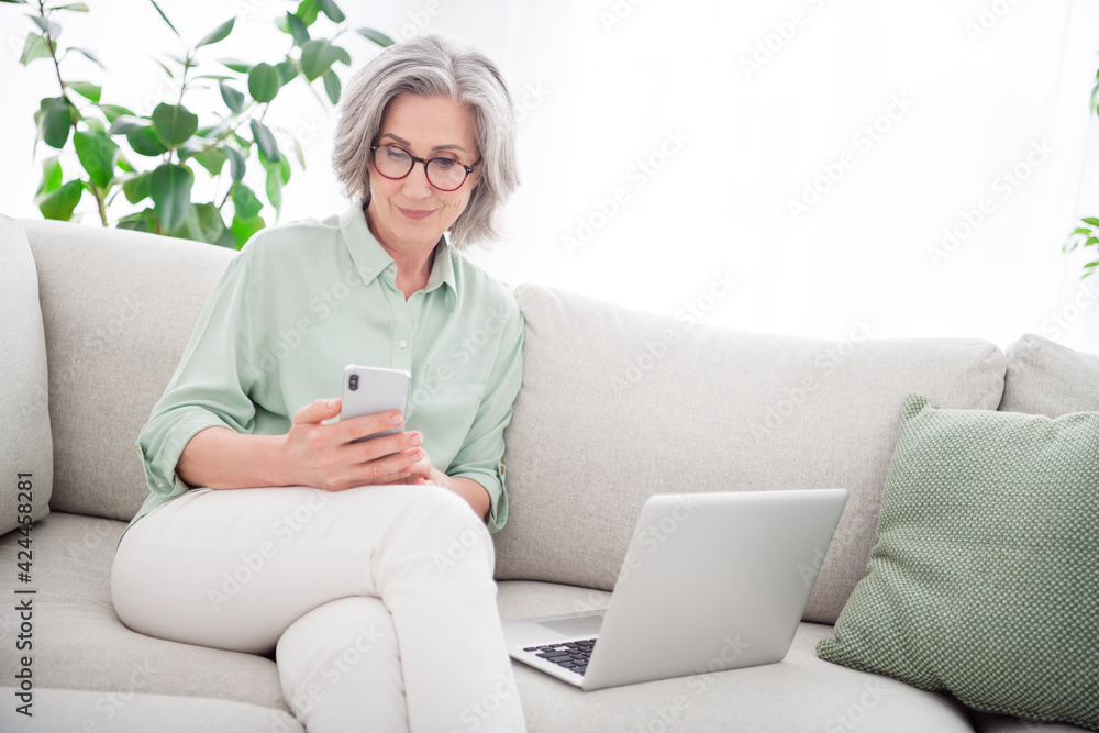 Portrait of pretty positive aged woman sit on sofa hands hold phone typing chatting spend free time home indoors Portrait of pretty positive aged woman sit on sofa hands hold phone typing chatting spend free time home indoors