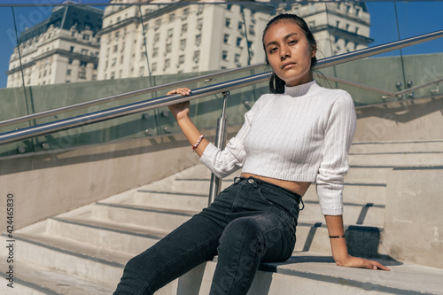 Young Latin woman sitting on the concrete staircase while enjoying the morning sun. Woman dressed in black jean and white top. © HC FOTOSTUDIO