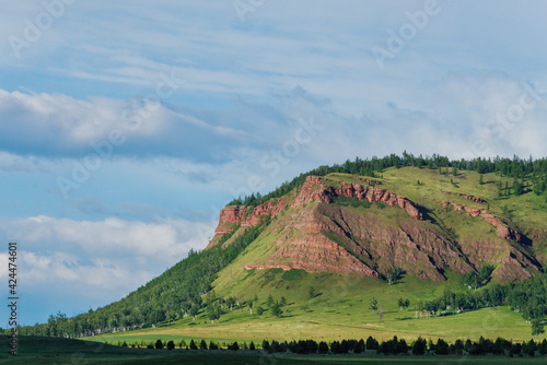 View of the red mountain in Khakassia weathering and erosion of rocks and red sandstone in the rays of the sun and the forest on the mountain