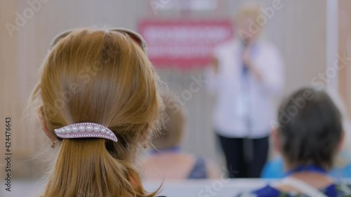 Woman listening to speech of unrecognizable lecturer at conference, seminar, meeting - back view, blurred background, view from auditorium. Education, communication, business concept