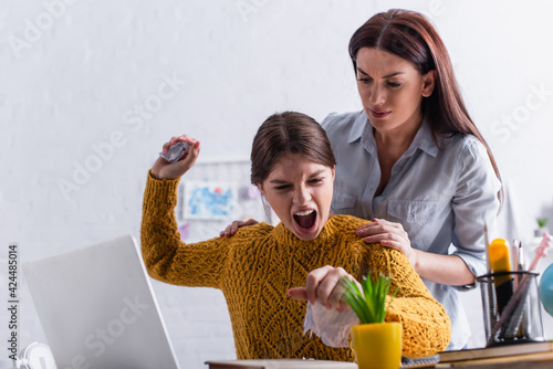 angry teenage girl tearing paper and screaming near worried mother while doing homework photo