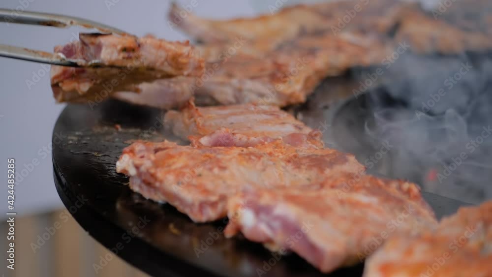 Chef holding tongs and grilling fresh juicy meat steaks on brazier with hot flame at summer local food market - close up view, slow motion. Outdoor cooking, gastronomy, cookery, street food concept Chef holding tongs and grilling fresh juicy meat steaks on brazier with hot flame at summer local food market - close up view, slow motion. Outdoor cooking, gastronomy, cookery, street food concept