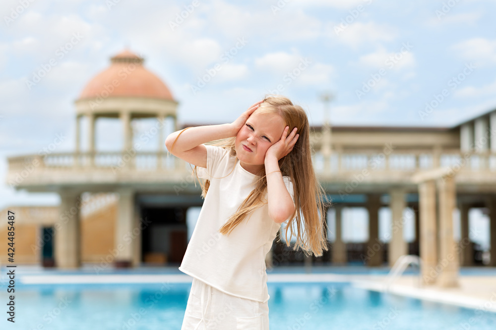 Fototapeta premium Emotions. Portrait of a little girl holding her head and showing her displeasure. Headache. In the background there is a building with a dome and a swimming pool