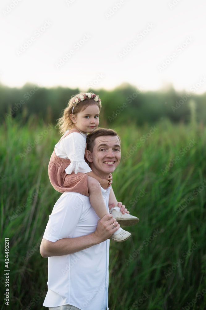 cute young dad with his daughter toddler, baby girl sitting on dad's neck, walking in the park in summer. Fatherhood concept. Soft focus and toning cute young dad with his daughter toddler, baby girl sitting on dad's neck, walking in the park in summer. Fatherhood concept. Soft focus and toning