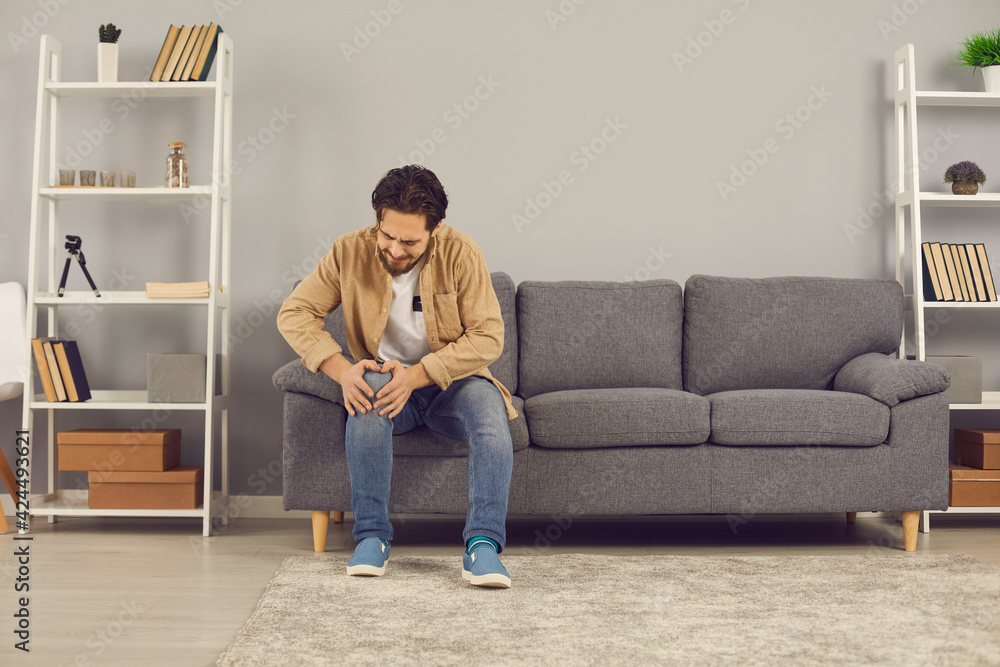 Unhappy man sitting on sofa at home and grimacing after hurting knee joint in domestic accident. Young guy, suffering from rheumatoid arthritis or osteoarthritis, feeling intense severe pain Unhappy man sitting on sofa at home and grimacing after hurting knee joint in domestic accident. Young guy, suffering from rheumatoid arthritis or osteoarthritis, feeling intense severe pain