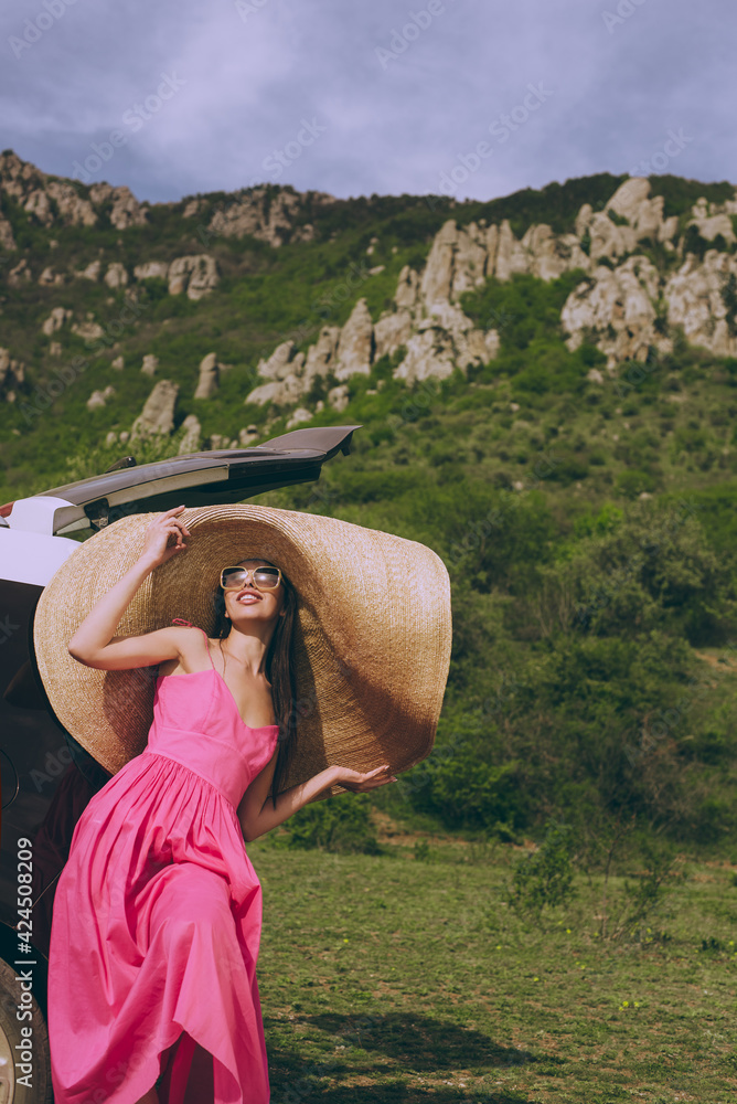 Young beautiful girl in a dress and a wide-brimmed hat near a hatchback car on a background of a mountain landscape.