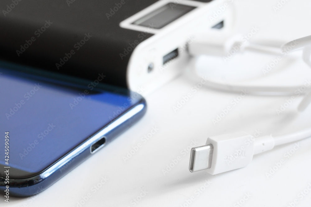 A large and capacious external battery - power bank lies next to the cable, usb type-c connector and a blue smartphone. The concept of recharging modern gadgets. Macro A large and capacious external battery - power bank lies next to the cable, usb type-c connector and a blue smartphone. The concept of recharging modern gadgets. Macro