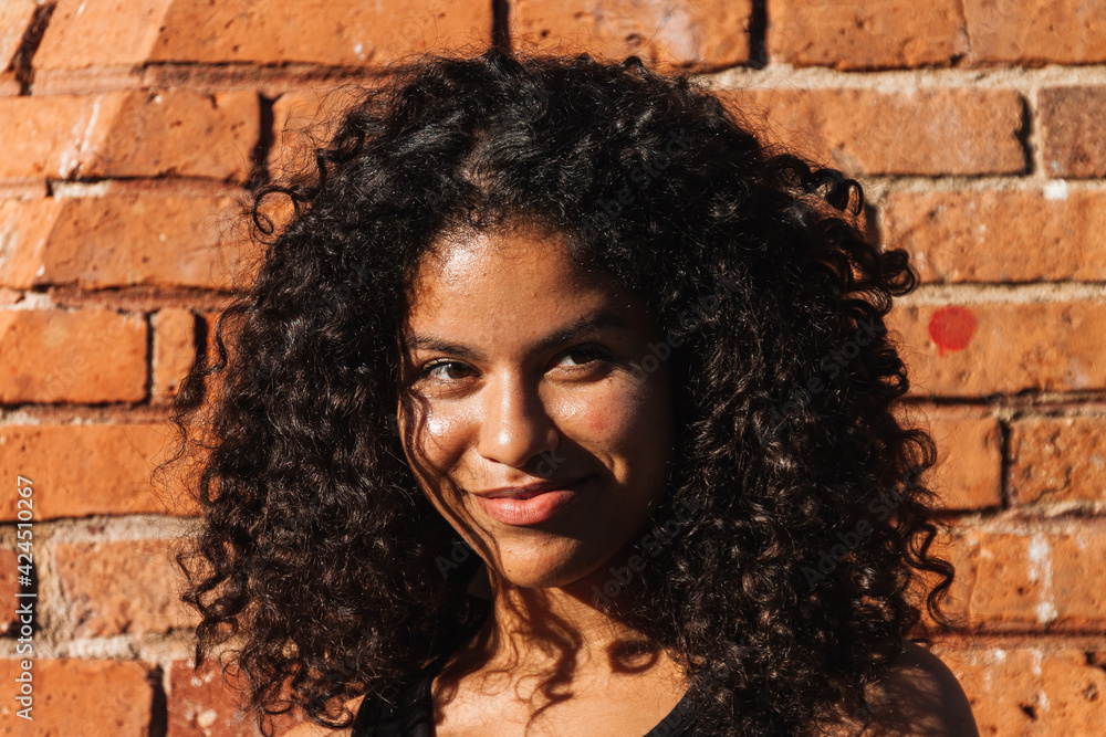 Woman with curly hair smiling while looking away. Brick wall on background Woman with curly hair smiling while looking away. Brick wall on background