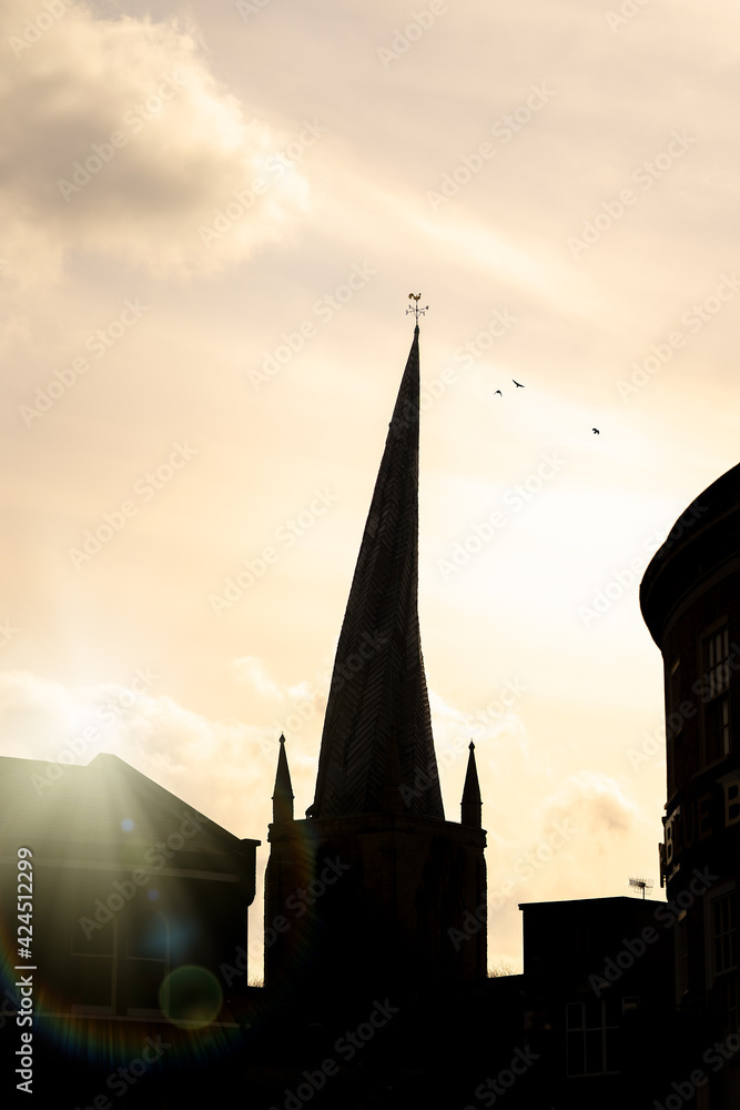Fototapeta premium Chesterfield Crooked Spire church silhouette of tower in town centre with sun setting behind and birds in sky cockerel weather vein on top. Historic St Marys parish worship building.