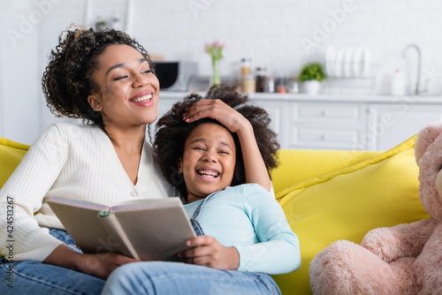 cheerful african american woman touching head of laughing daughter while sitting on sofa with book