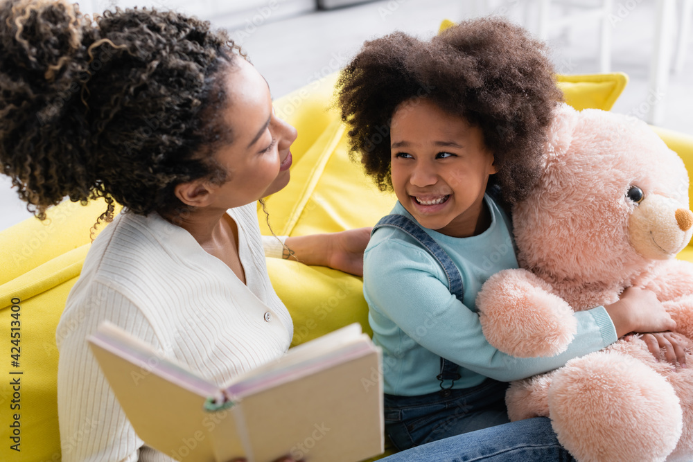 happy african american girl holding teddy bear near mother with book on blurred foreground happy african american girl holding teddy bear near mother with book on blurred foreground