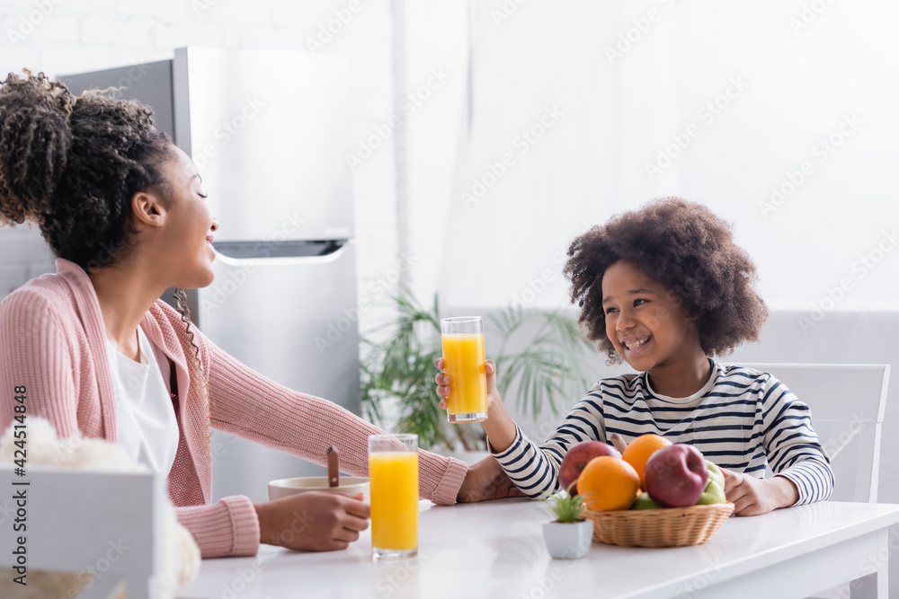 joyful african american girl holding glass of orange juice near mother during breakfast