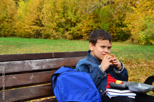 Close-up of a small schoolboy sitting on a Park bench and opening his school backpack during a lunch break. eating a sandwich out of a lunch box