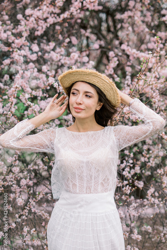 Beautiful young caucasian brunette woman in luxury long dress among the blossoming sakura flowers. enjoying spring Early spring concept. Rest on nature vacation. Girl in straw hat.