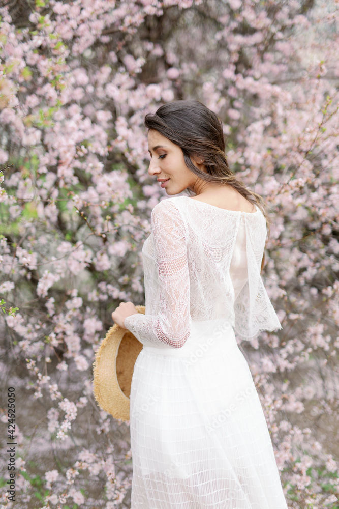 Beautiful young caucasian brunette woman in luxury long dress among the blossoming sakura flowers. enjoying spring Early spring concept. Rest on nature vacation. Girl in straw hat. Beautiful young caucasian brunette woman in luxury long dress among the blossoming sakura flowers. enjoying spring Early spring concept. Rest on nature vacation. Girl in straw hat.