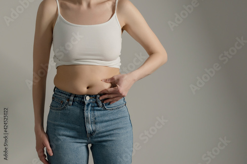 Woman in jeans and white shirt pinching her belly fat. Woman`s hips closeup  raw studio shot in grey background. Dieting and fat loss concept. Stock  Photo