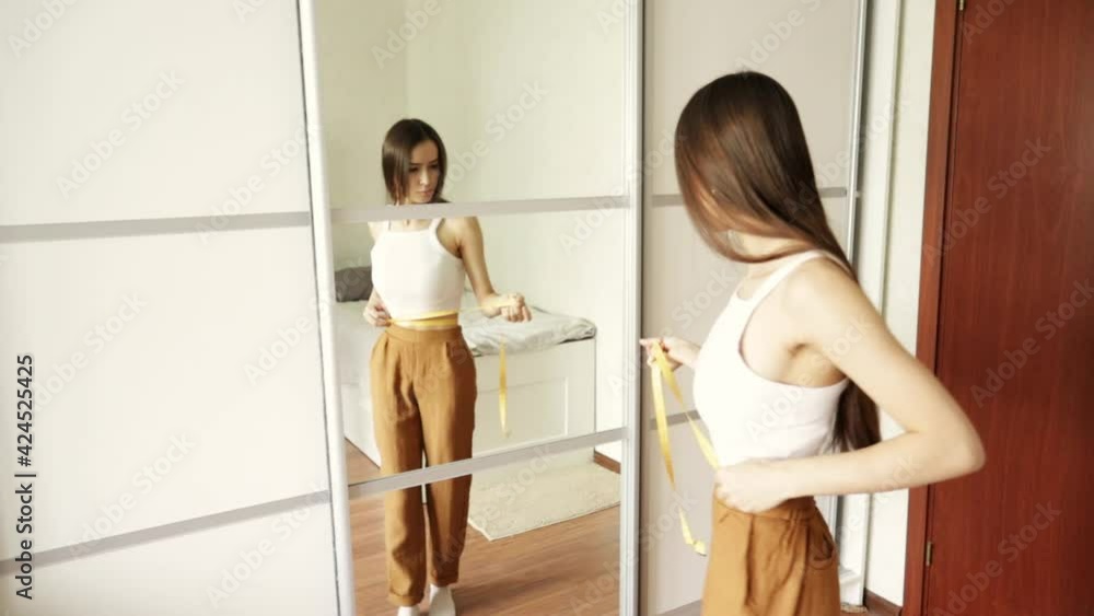Body care, beauty, healthy eating, teen girl in front of the mirror measures her waist with a ribbon Body care, beauty, healthy eating, teen girl in front of the mirror measures her waist with a ribbon