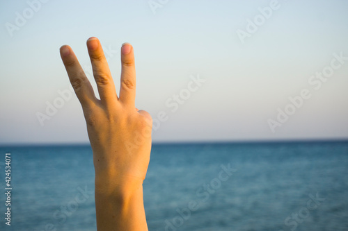 Hand doing showing number three gesture symbol on blue summer sky nature background. Gesturing number 3. Number three in sign language. Third, counting down three concept. Three fingers up. 