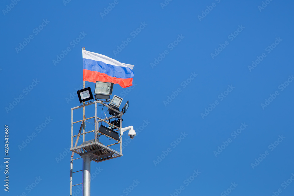 A tower with spotlights and a video surveillance camera. The flag of the Russian Federation develops against a blue sky. A tower with spotlights and a video surveillance camera. The flag of the Russian Federation develops against a blue sky.
