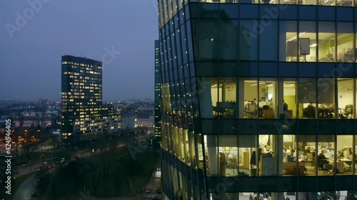 Drone shot at the office windows of a skyscraper at night, the camera moves from bottom to top panning the office windows and partially the city of Warsaw in Poland.