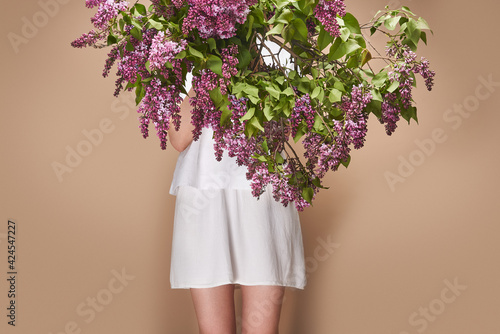 Faceless woman in white clothes holds the bouquet of lilac flowers on the beige background indoors